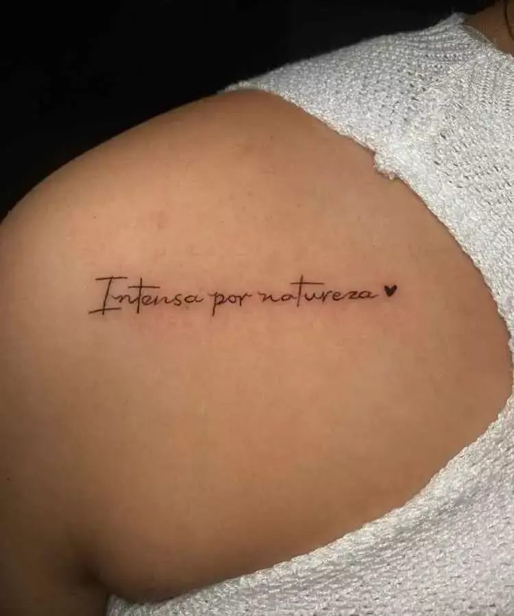 A imagem apresenta uma tatuagem no ombro com a frase delicada "Intensa por natureza" escrita em uma fonte fina e cursiva, acompanhada de um pequeno coração ao lado. A pele está coberta parcialmente por uma blusa branca de tecido tricotado.