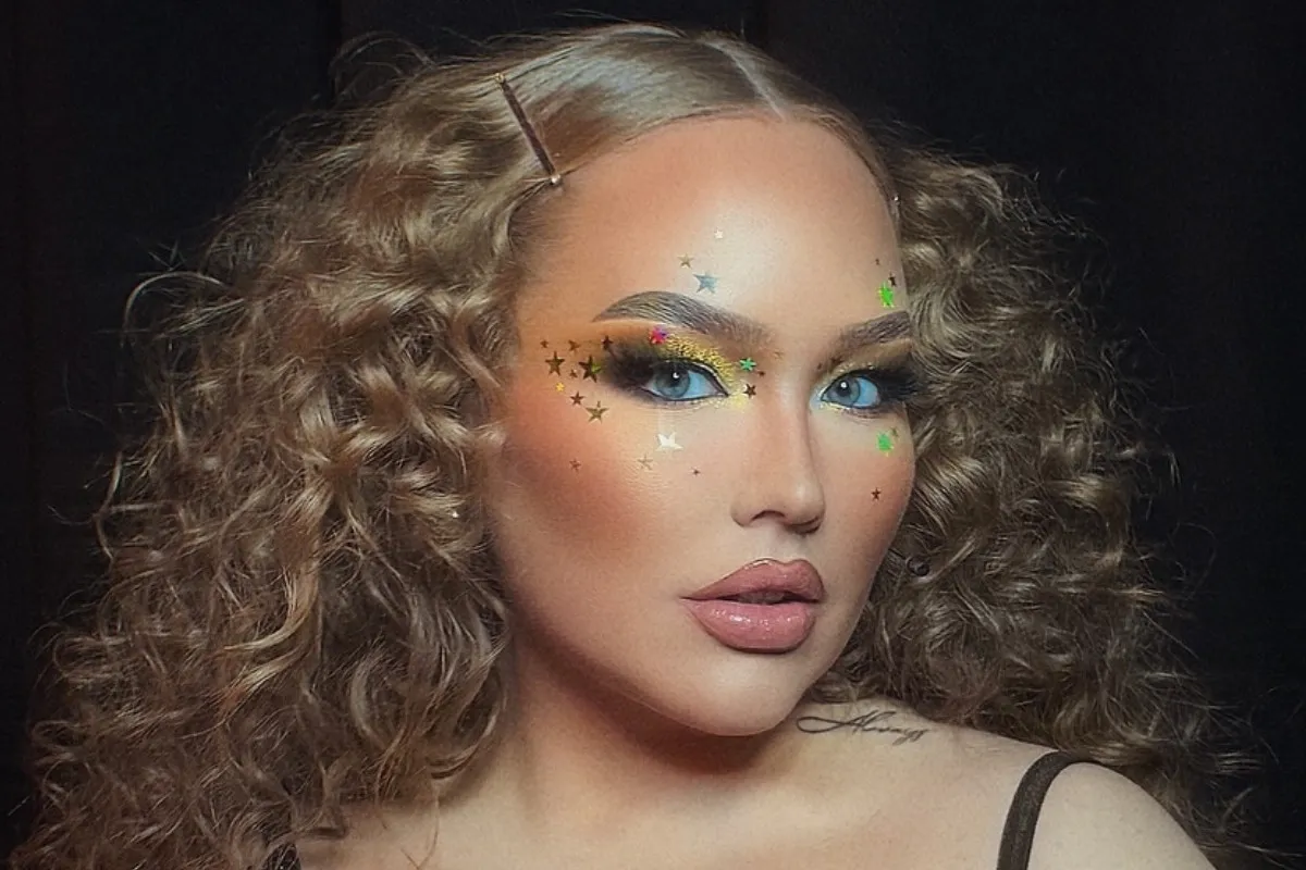 Close-up de nikkietutorials com maquiagem artística em fundo escuro. Seus olhos são azuis, com sombra metálica; os lábios, rosa-nude brilhantes. Pequenos decalques de estrelas adornam suas bochechas. Cabelos castanhos encaracolados, com grampos metálicos.