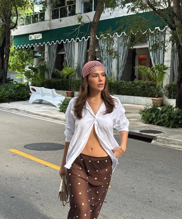 Mulher de cabelo castanho em uma rua, usando um chapéu de crochê rosa, uma camisa social branca aberta, mostrando o abdômen, e calças transparentes de bolinhas marrons sobre um biquíni.
