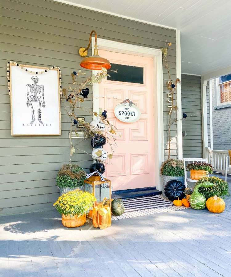 Porta de entrada cinza e pêssego decorada com galhos secos, corvos, abóboras, quadro de esqueleto e flores 