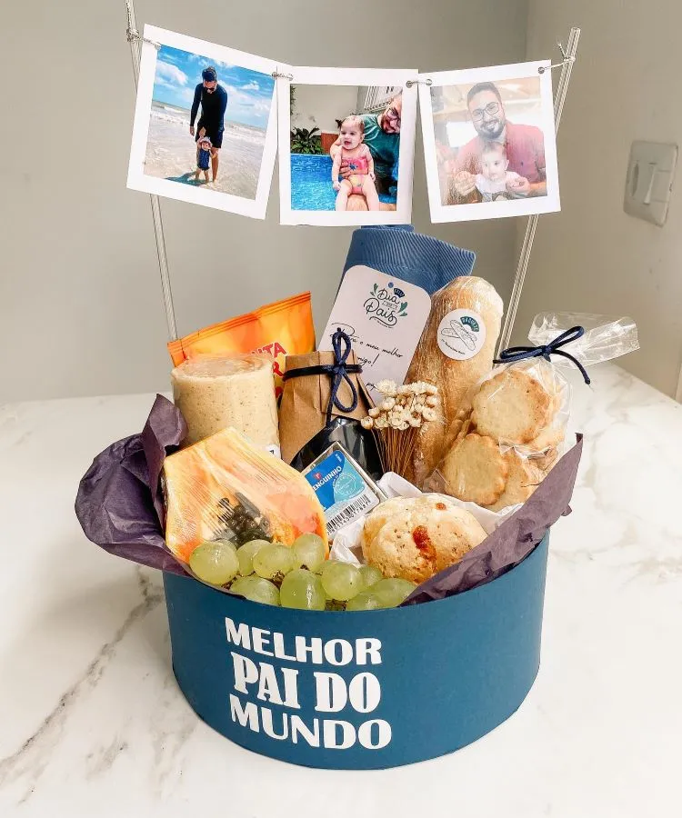 A imagem mostra uma cesta de Dia dos Pais com itens gourmet, incluindo queijos, pães, biscoitos e frutas. Acima, fotos penduradas em um fio com prendedores exibem momentos felizes. A cesta tem a frase “MELHOR PAI DO MUNDO” em destaque na frente.

