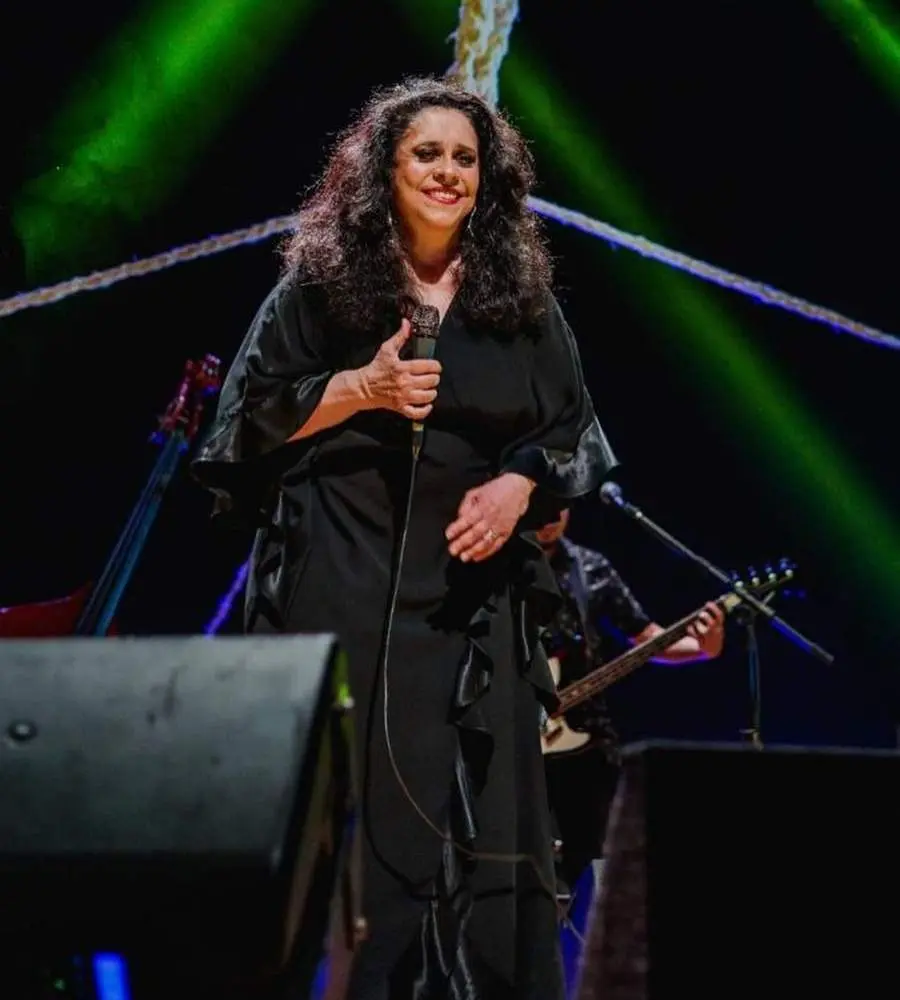 Gal Costa em palco cantando com microfone nas mãos. No fundo, músicos com seus instrumentos musicais.
