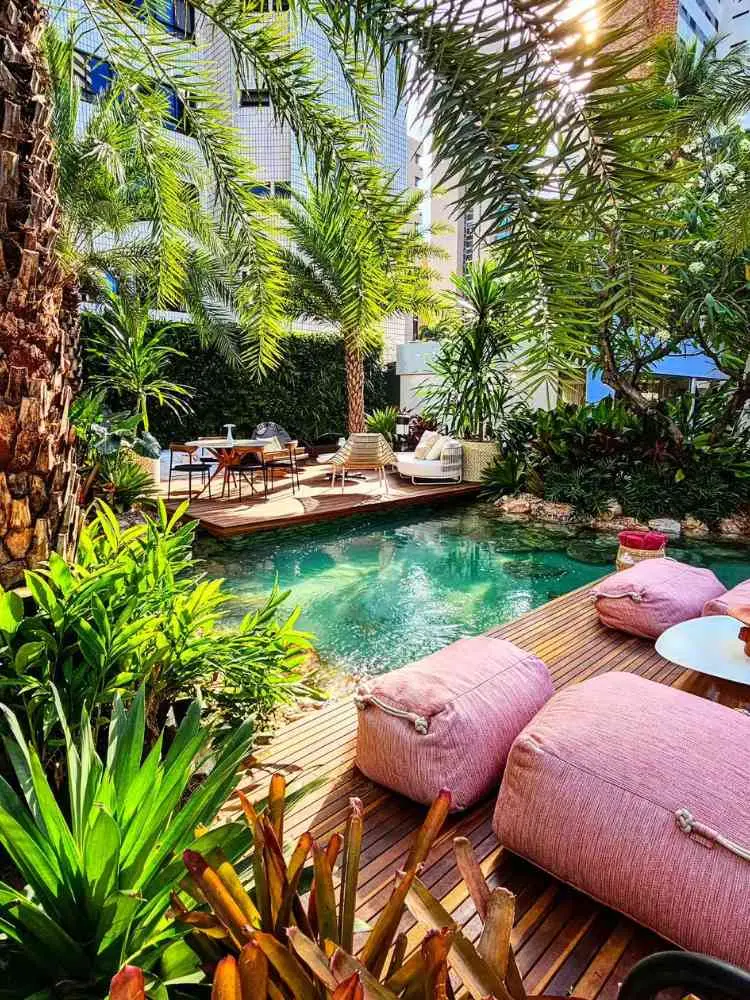 Quintal com piscina biológica, muitas árvores em volta e estofados na cor rosa. 
