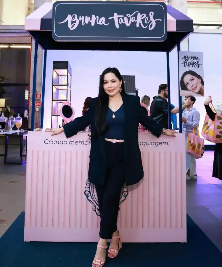 Bruna Tavares posa em frente a um estande decorativo da marca Bruna Tavares, com elementos em rosa e azul escuro. Ela veste blazer e calça preta, sapatos rosa, e sorri. Ao fundo, pessoas exploram o ambiente iluminado.