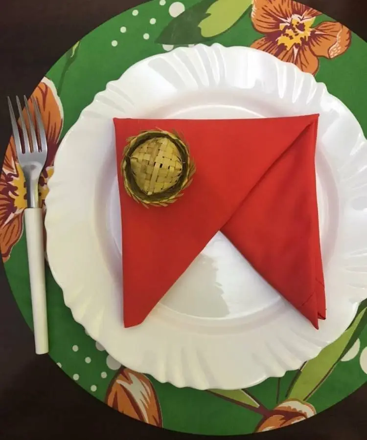 Uma mesa posta com um prato branco sobre um sousplat verde floral. Um guardanapo vermelho está dobrado em forma de bandeira e preso com um mini chapéu de palha.