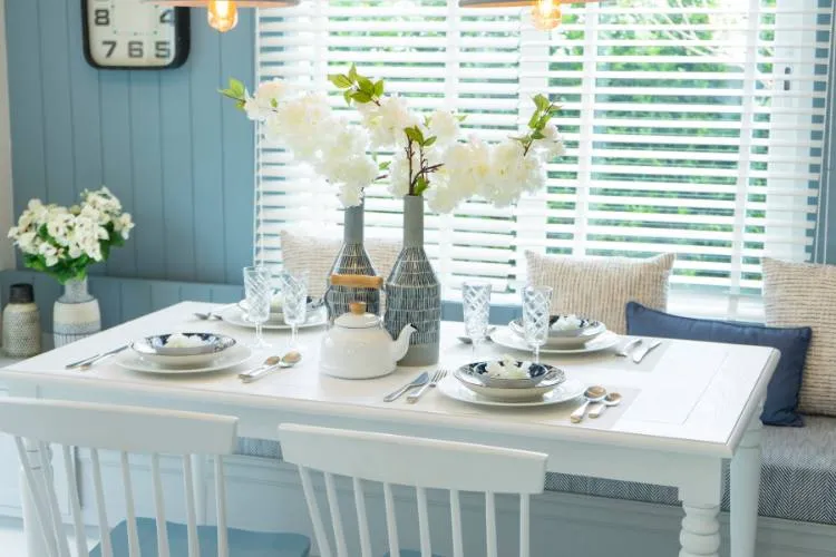 A imagem mostra uma mesa branca e retangular. A mesa está servida com pratos brancos com padrões florais azuis, guardanapos brancos e talheres de prata. O centro da mesa é um vaso com flores brancas e um bule branco. O fundo consiste em uma parede azul com um relógio e uma janela com persianas brancas. O fundo também inclui um banco com almofadas azuis e brancas. A imagem transmite uma sensação de simplicidade e conforto.