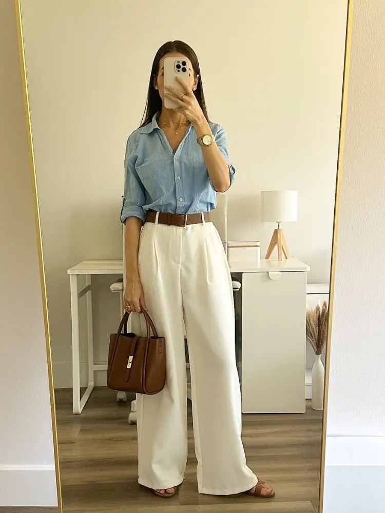 Mulher de pele clara tirando foto no espelho usando camisa azul e calça social clara 
