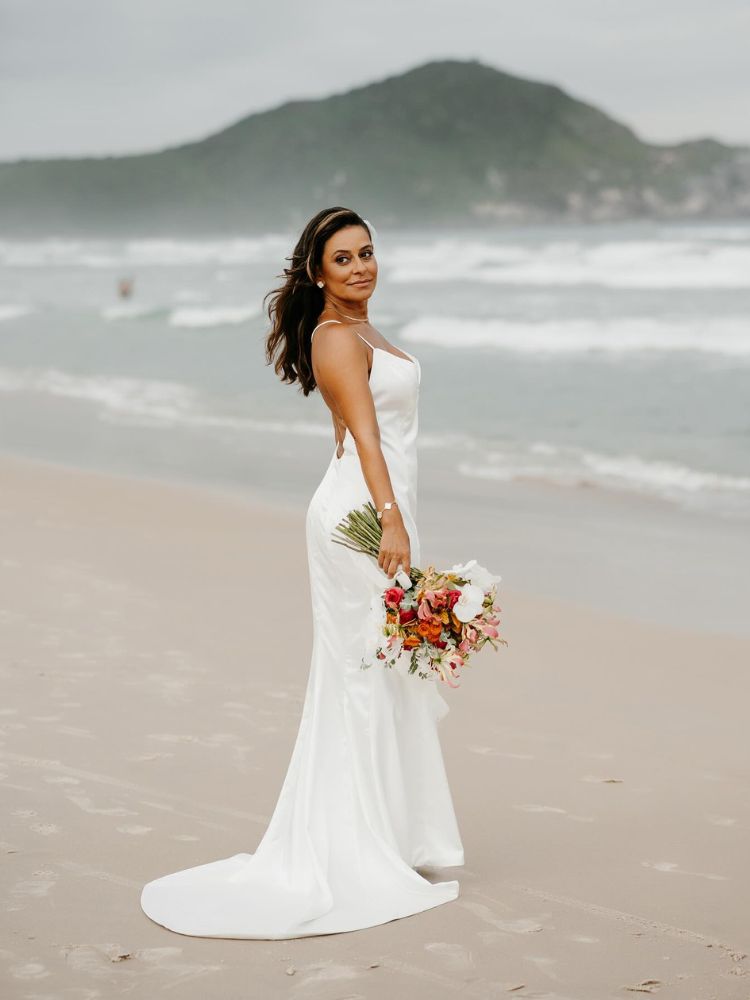 Noiva de pele morena, cabelo solto, vestido de seda simples e buquê