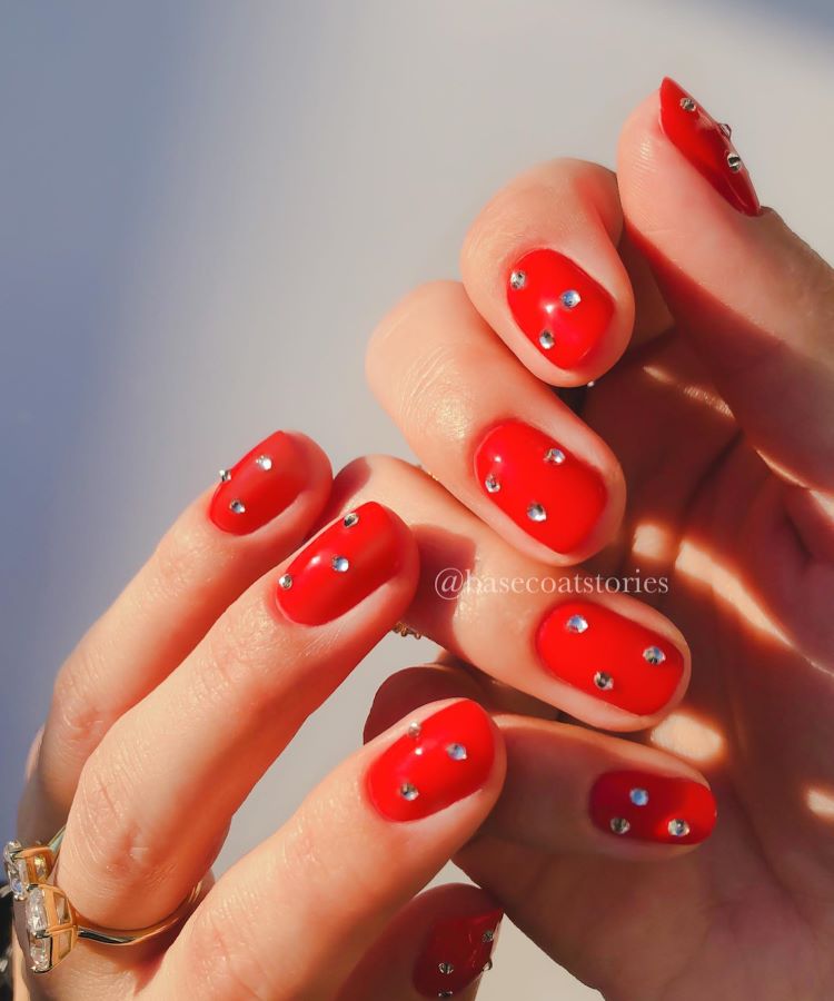 A imagem mostra mãos com unhas pintadas de vermelho vibrante, decoradas com pequenas pedras prateadas, criando um efeito brilhante e elegante. As mãos estão iluminadas por luz natural, destacando o brilho do esmalte e das pedras. Há um anel dourado com um design moderno em um dos dedos. O fundo é neutro, permitindo que o foco permaneça nas unhas e nos detalhes da decoração.