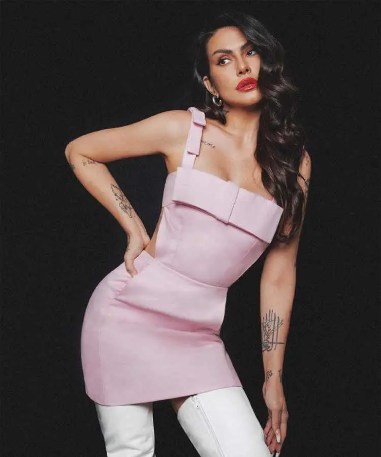 Cleo Pires em fundo preto posando, está vestindo vestido rosa de alças e bota alta branca. Ela possui tatuagem nos dois braços e pulsos