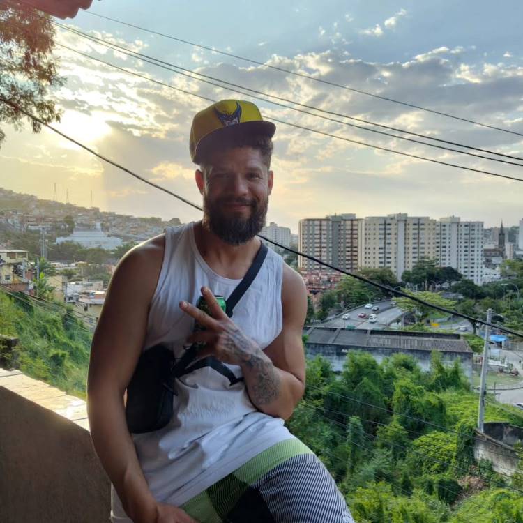Juninho de regata branca, bermuda e boné sentado na mureta com vista para árvores e prédios.
