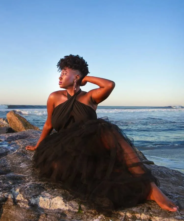 Uma mulher sentada em uma rocha na praia, com o oceano ao fundo. Ela usa um vestido preto esvoaçante e tem um corte de cabelo cacheado curto e raspado nas laterais, olhando para o lado.