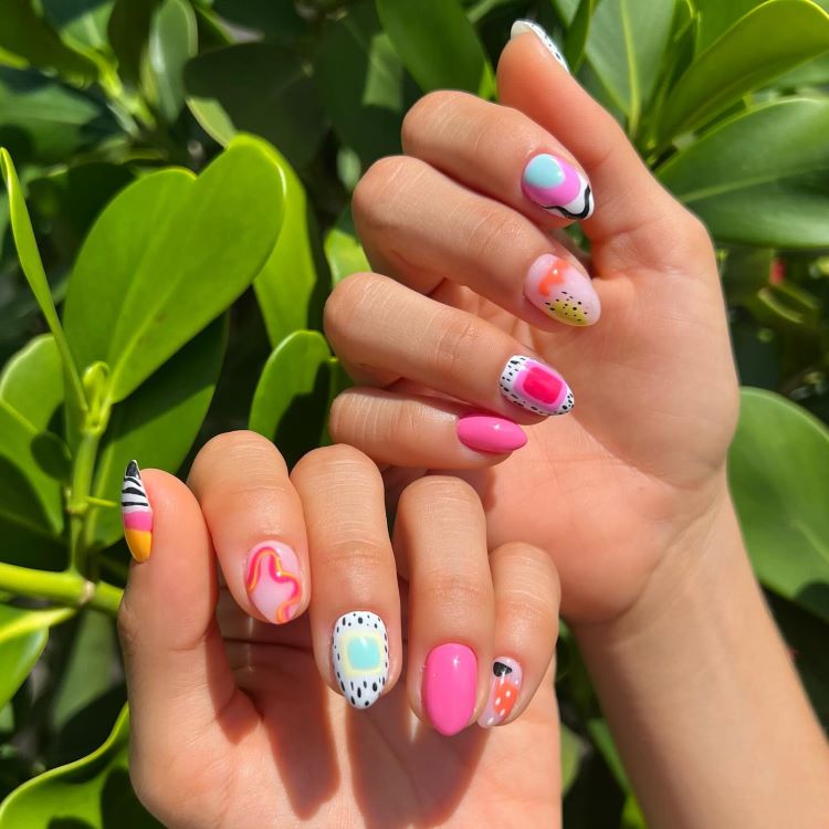 Mãos exibindo unhas decoradas com designs coloridos e abstratos, incluindo frutas tropicais, padrões geométricos, linhas onduladas e detalhes pontilhados. Tons vibrantes de rosa, laranja, amarelo e azul destacam-se sob luz solar intensa. Fundo natural com folhas verdes desfocadas.