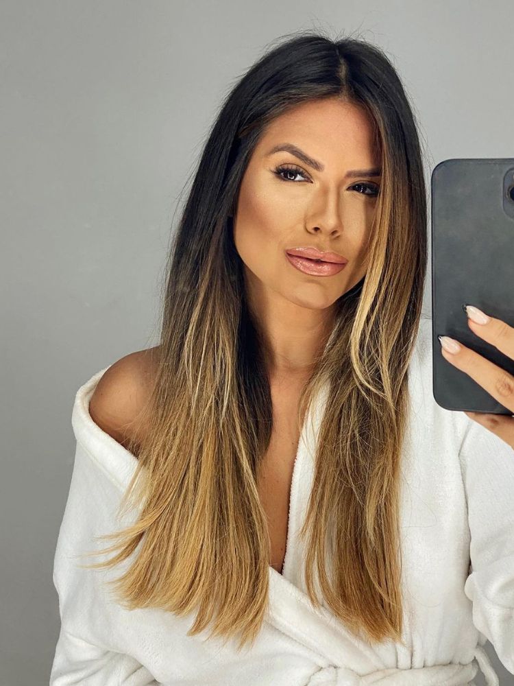 Foto de Carolina, participante do Puxadinho do BBB 24. Ela segura o celular em frente ao espelho e usa um roupão branco