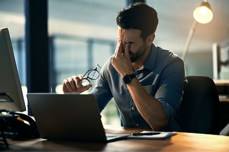 A imagem mostra um homem sentado em uma mesa de trabalho, aparentando cansaço. Ele segura seus óculos em uma mão enquanto pressiona os olhos com a outra. Em frente, há um laptop e luz suave no ambiente corporativo.





