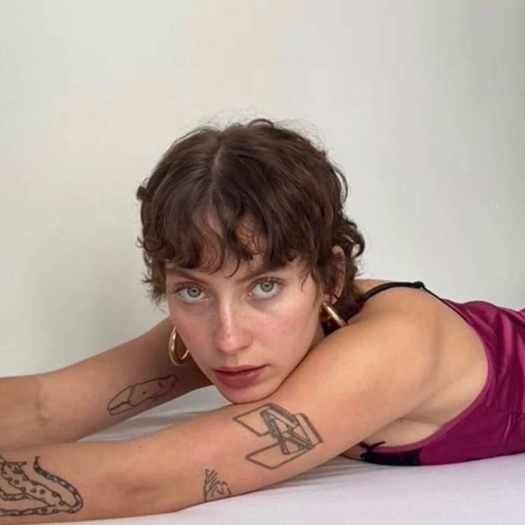 Mulher com cabelo castanho curto estilo messy, franja leve na testa e expressão séria. Pele clara, olhos verdes destacados e batom bordô. Usa blusa de alça vinho, brincos de argola dourados, exibindo tatuagens geométricas e orgânicas nos braços.