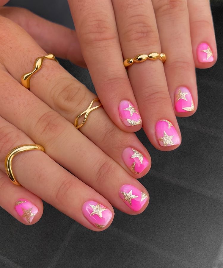 Mãos com unhas pintadas em tom rosa vibrante, decoradas com estrelas douradas brilhantes e detalhes em glitter. Dedos adornados com anéis dourados de designs variados. Fundo cinza escuro desfocado, destacando o visual elegante e moderno das unhas.