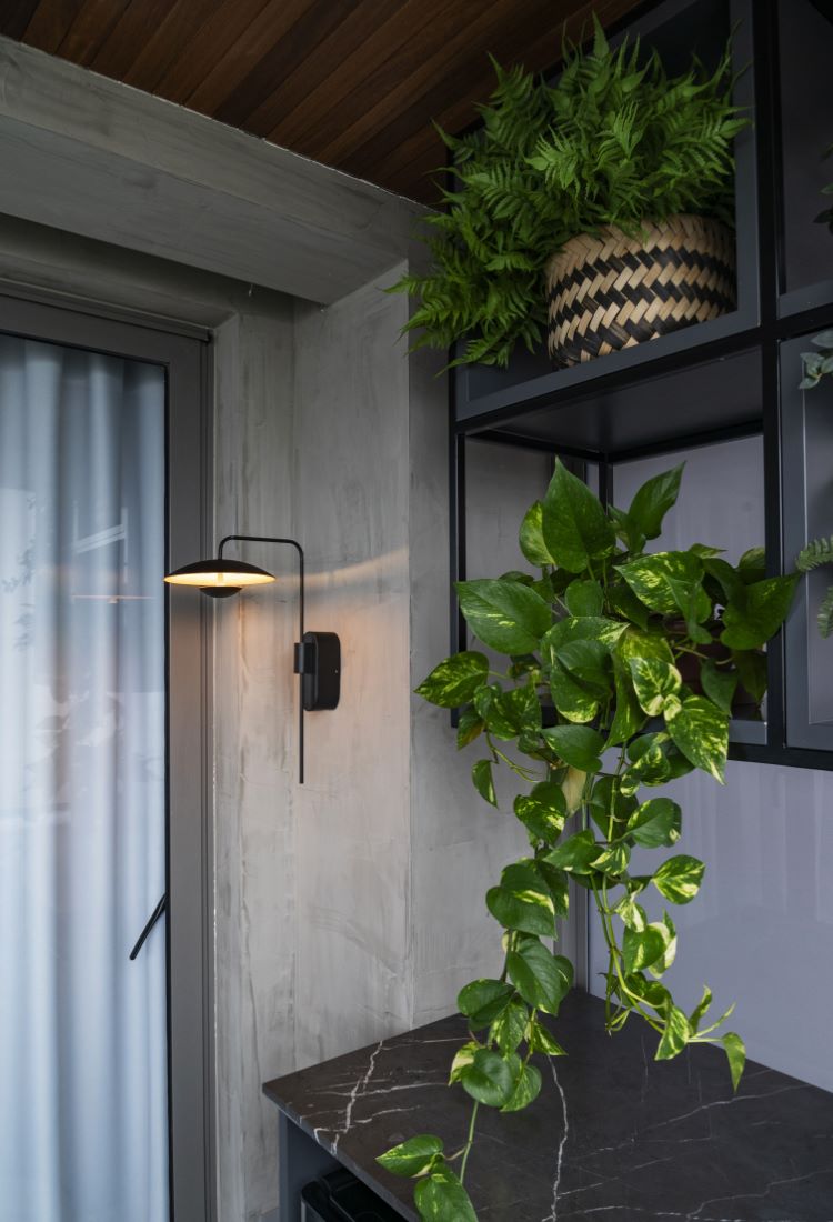 A imagem mostra um canto de varanda decorado com plantas, destacando uma prateleira preta com vasos de samambaia e uma planta pendente. A parede de cimento queimado e o teto de madeira trazem um toque moderno ao ambiente, enquanto uma luminária de parede em preto adiciona iluminação suave. As plantas verdes contrastam com o tom neutro da parede, criando um ambiente natural e agradável. Esse espaço é um ótimo exemplo de como decorar apartamento com plantas, utilizando prateleiras e vasos suspensos para otimizar o espaço e trazer vida ao ambiente. A bancada de mármore escuro complementa o visual sofisticado e funcional.


