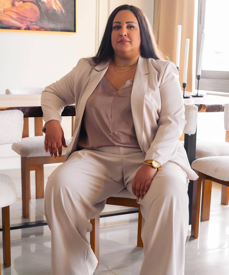 Mulher sentada em cadeira em sala elegante, veste terno bege e blusa de cetim. Usa colares dourados e relógio. Fundo com mesa de madeira, cadeiras estofadas brancas e obras de arte ao lado de janelas.






