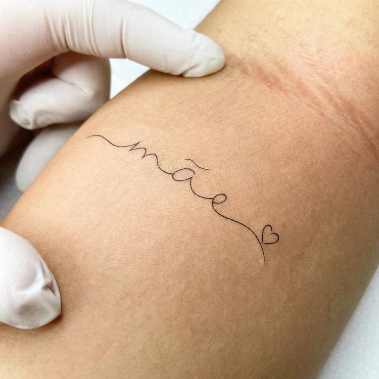 A imagem mostra o antebraço de uma pessoa com uma tatuagem fina e delicada da palavra "mãe" em escrita cursiva, seguida por um pequeno coração, ambos em preto. A pele apresenta algumas marcas e uma mão enluvada de branco toca a pele.







