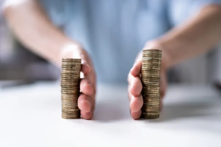 Duas pilhas de moedas douradas em uma superfície branca, com mãos próximas separando-as cuidadosamente, simbolizando gestão financeira, controle de gastos e estratégias para organização de recursos durante momentos de planejamento ou crise econômica. Fundo levemente desfocado.
