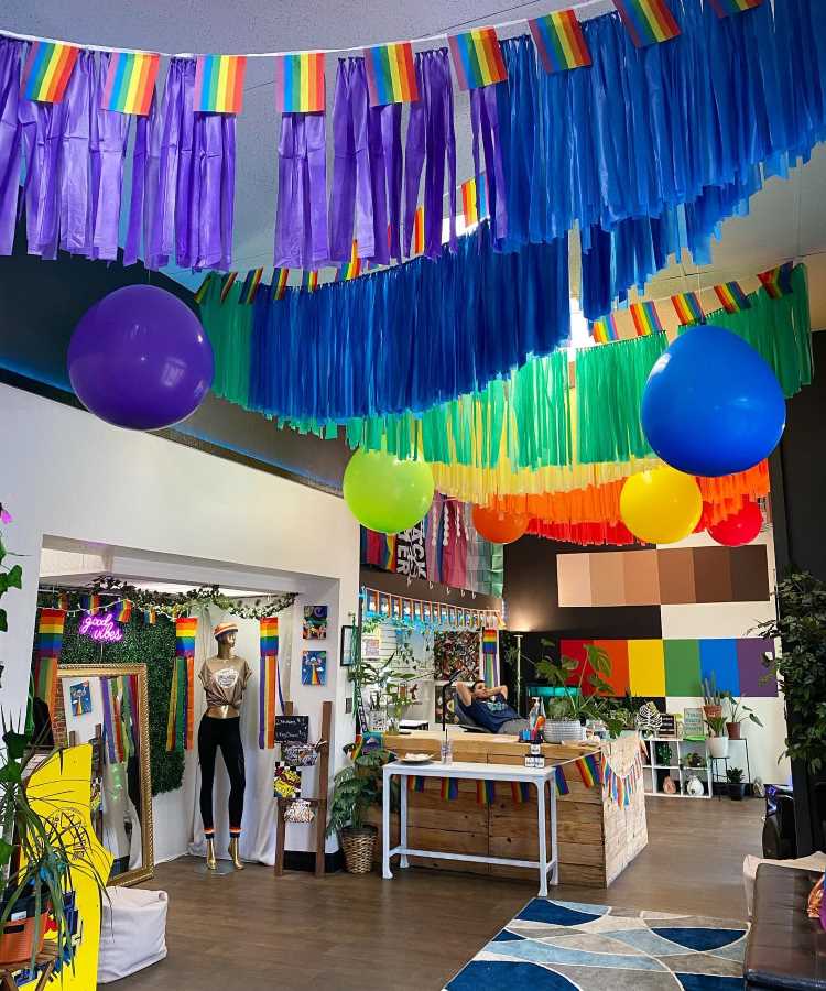 Espaço decorado com bandeiras arco-íris, fitas coloridas e balões vibrantes pendurados no teto. Manequim com roupa estilosa ao lado de plantas e espelho. Balcão rústico com decoração temática e sofá ao fundo criam ambiente acolhedor e inclusivo.