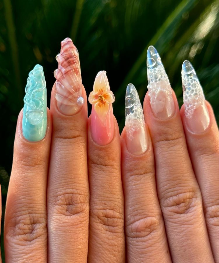 A imagem apresenta unhas longas, decoradas com nail art temática "seashell nails". Cada unha tem design único, combinando tons pastel, efeito furta-cor, textura ondulada, pérolas, bolhas e uma flor delicada. O visual é artístico e inspirado no oceano.
