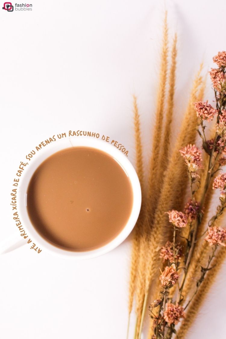 Uma xícara branca cheia de café com leite está no centro de uma superfície branca. Ao lado, um buquê de grama seca e flores desidratadas de tom rosado. Na borda da xícara, a frase em português: "Até a primeira xícara de café, sou apenas um rascunho de pessoa".