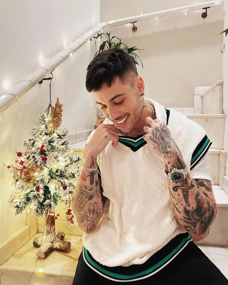 Gui Araújo sentado em escada ao lado de uma mini árvore de natal, veste colete de tricô branco e calça preta. Ele possui tatuagens nos braços, pulsos e pescoço