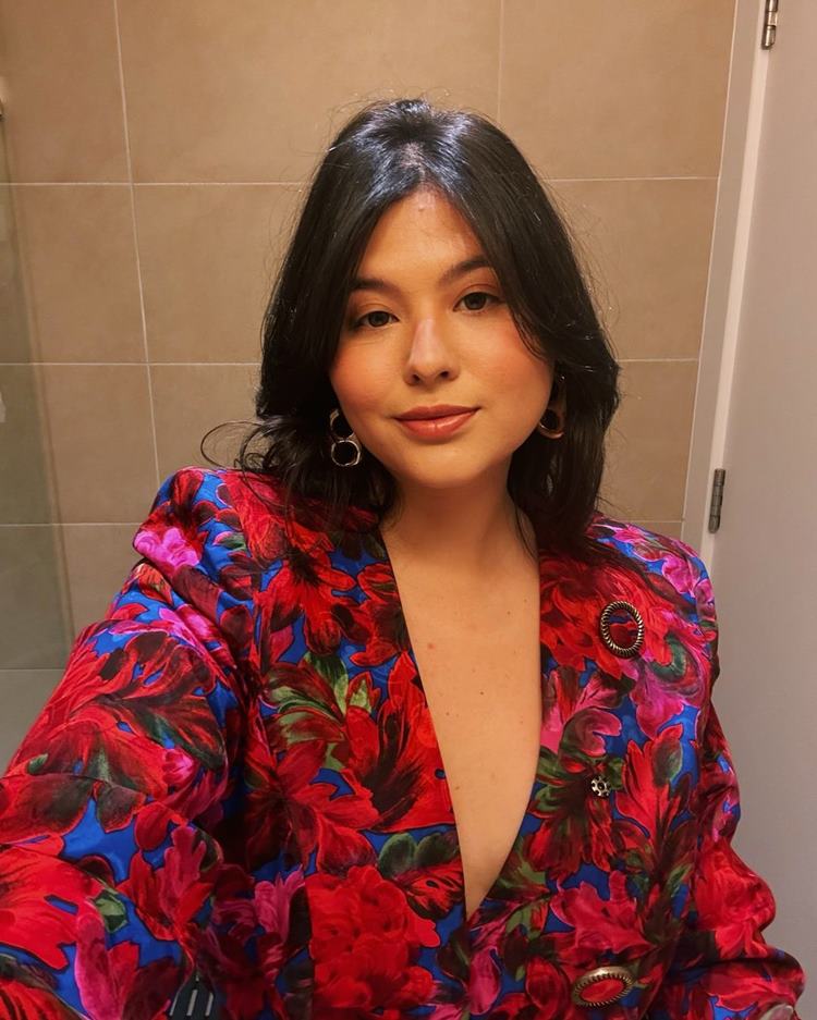 Mulher com cabelos escuros e soltos, usando blazer floral vermelho e azul com decote profundo. Ela está em um ambiente com azulejos bege, olhando diretamente para a câmera, com expressão serena. Usa brincos de argola dourada, em um estilo elegante. Ela é uma das meninas da lista de blogueiros CLTs.


