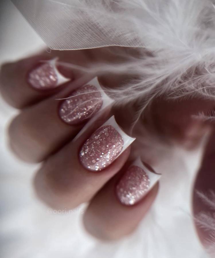 Unhas curtas em formato quadrado com esmalte rosa glitter e pontas francesas brancas destacando sofisticação. Detalhes delicados de plumas brancas criam uma atmosfera suave e romântica. O brilho do glitter contrasta elegantemente com o fundo desfocado etéreo.





