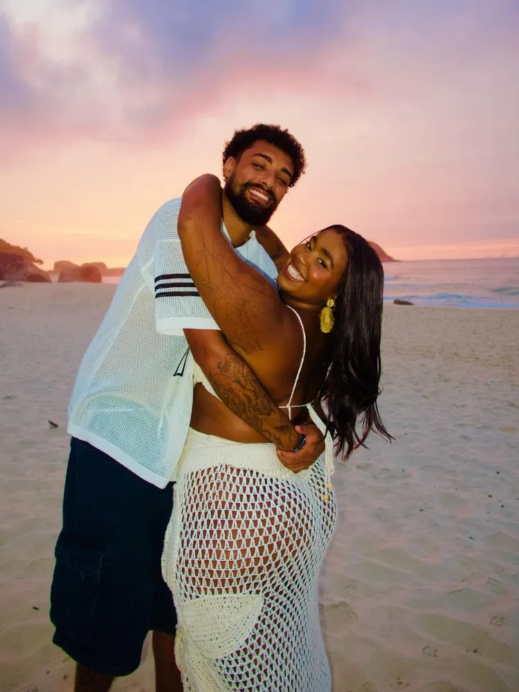 João Lemos de blusa branca e calça preta abraçado na praia com Mari Lobo, de biquíni e saia de croche brancos