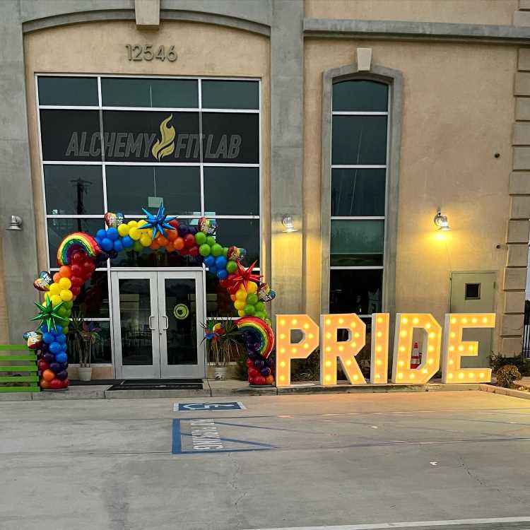 Fachada do Alchemy Fit Lab decorada com arco de balões arco-íris vibrantes, estrelas coloridas e dois painéis com arco-íris. Letras iluminadas formam a palavra "PRIDE". Ambiente celebra o Orgulho LGBTQIA+, com atmosfera festiva e acolhedora ao entardecer.
