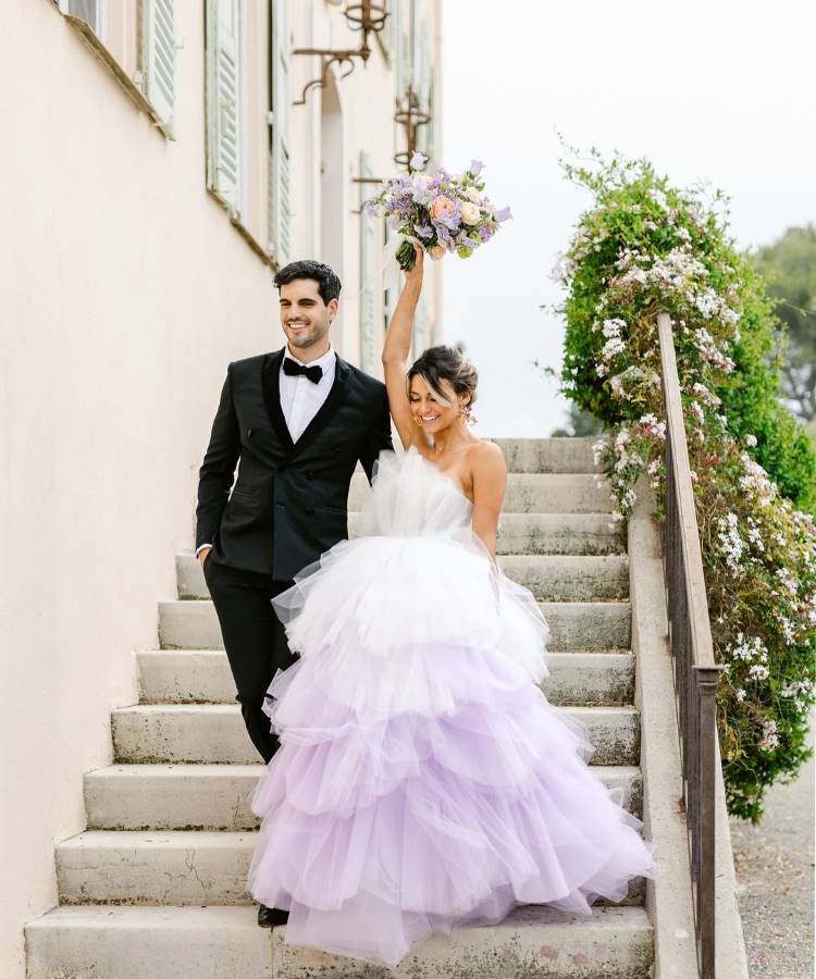 Um casal de noivos descendo uma escadaria de pedra. A noiva, sorrindo e com braço erguido segurando um buquê, veste um volumoso vestido de noiva branco e lilás. O noivo veste um elegante terno preto.







