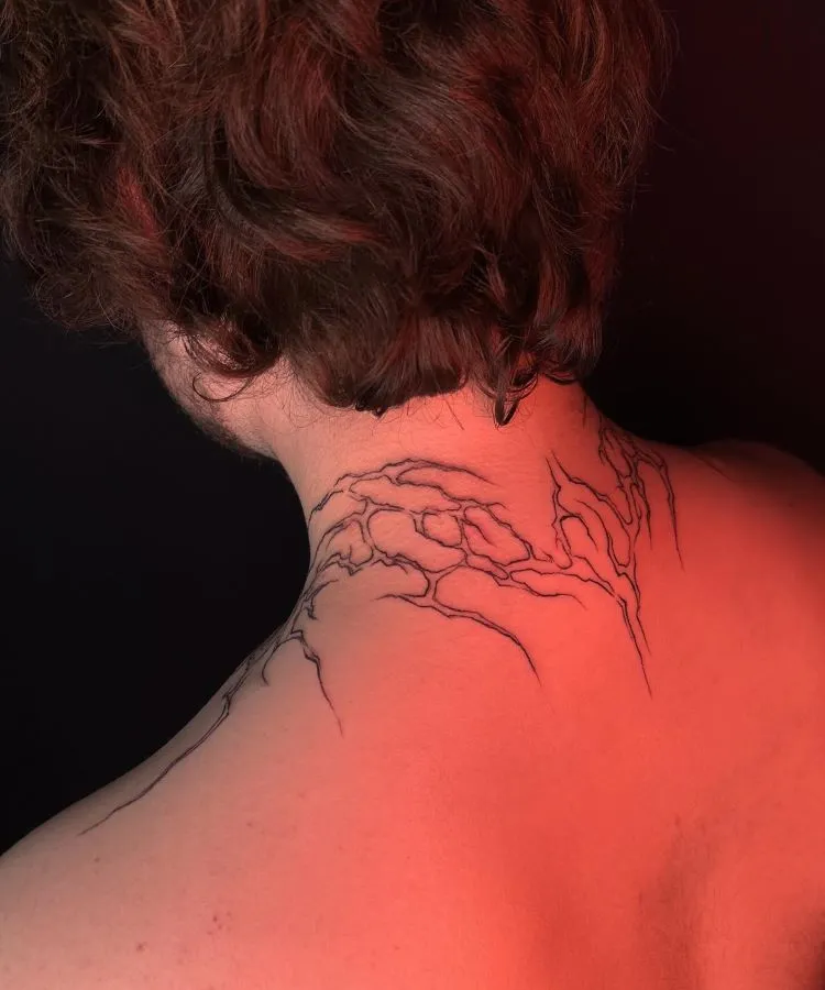 A imagem mostra uma pessoa de costas com cabelo castanho ondulado. Ela tem uma tatuagem no pescoço de galhos finos e detalhados, desenhados em linhas pretas que se estendem da base do crânio até os ombros. A tatuagem é delicada e parece parte de uma árvore.




