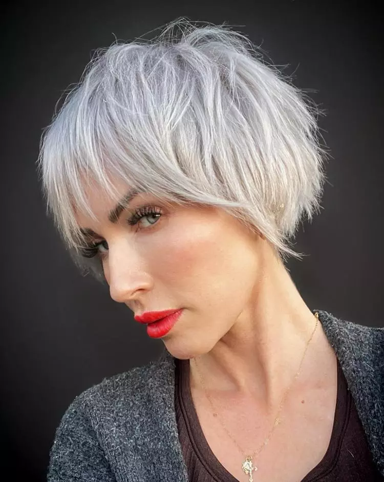 Mulher com corte de cabelo curto bixie cinza, usando batom vermelho, tem olhos claros, e está usando roupa de lã