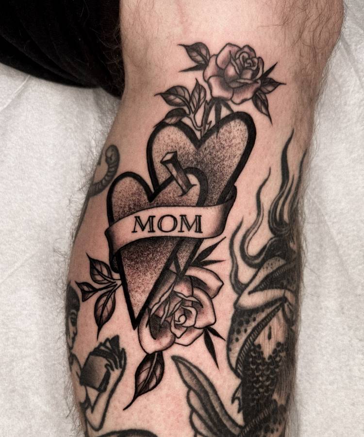 A imagem exibe uma tatuagem no estilo "old school" na perna de uma pessoa. Ela apresenta dois corações texturizados, um deles transpassado por um prego, e uma faixa com a palavra "MOM". Rosas com folhas adornam a parte superior e inferior. Há outras tatuagens visíveis na mesma perna.







