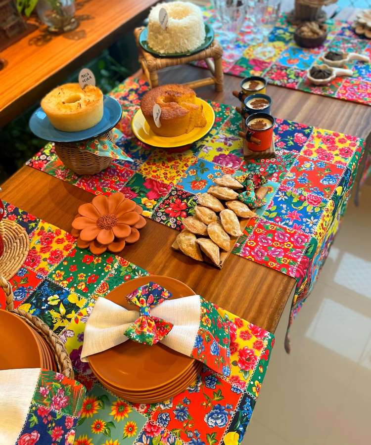 Uma mesa ricamente decorada para festa junina com uma toalha de retalhos coloridos, espigas de milho adornadas com fita xadrez, bolo de milho e canecas esmaltadas, criando um ambiente acolhedor e festivo.