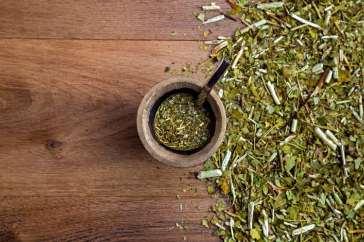 Erva-mate espalhada em superfície de madeira com cuida de chimarrão ao lado