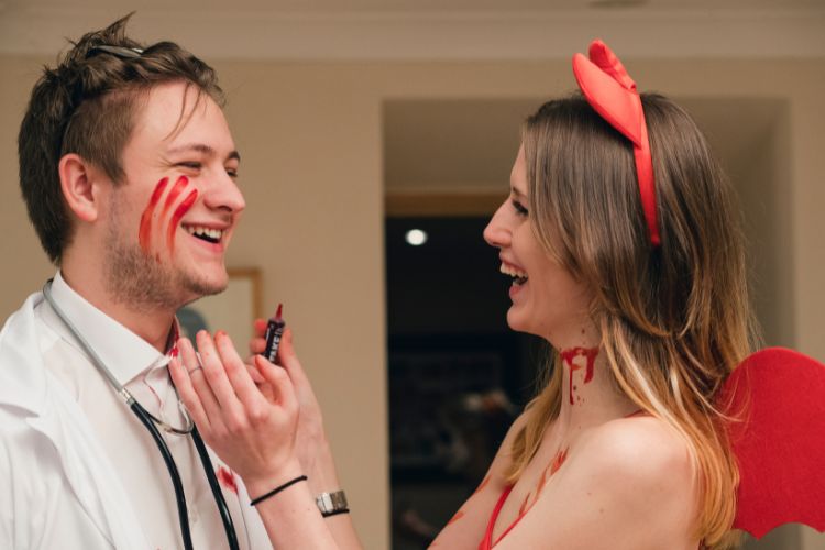 Mulher fantasiada de diabinha com sangue no pescoço rindo e pintando rosto de homem fantasiado de médico com sangue falso
