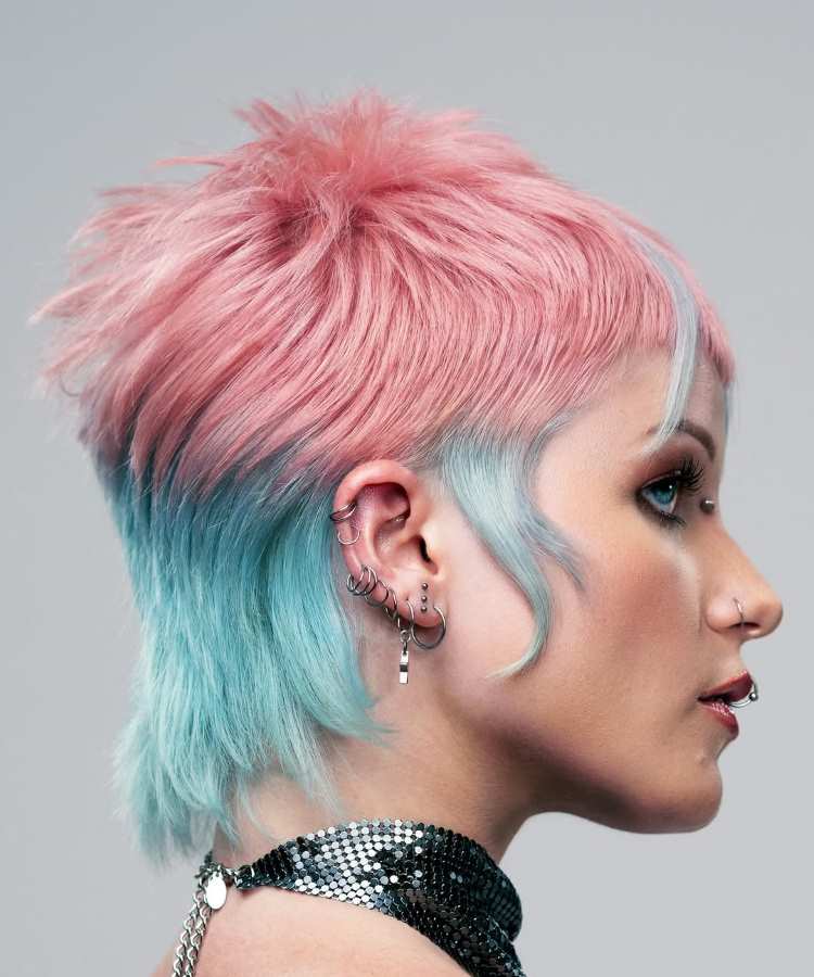 Mulher com cabelo estilo mullet em tons rosa pastel e azul, com franja curta. Usa vários piercings nas orelhas, piercing no nariz, maquiagem com delineado marcante e batom vermelho. Veste gargantilha brilhante em fundo neutro iluminado.