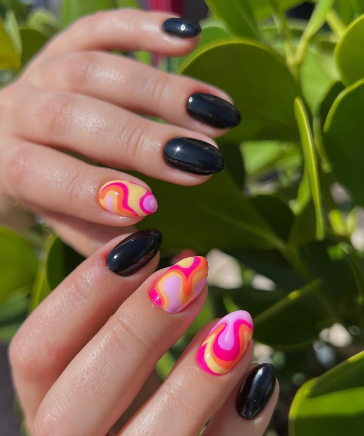 Mãos com unhas decoradas em esmalte preto brilhante e detalhes psicodélicos em cores vibrantes, como rosa, laranja e amarelo, formando padrões ondulados. Fundo com folhas verdes desfocadas, iluminado por luz natural, criando um contraste tropical e moderno.