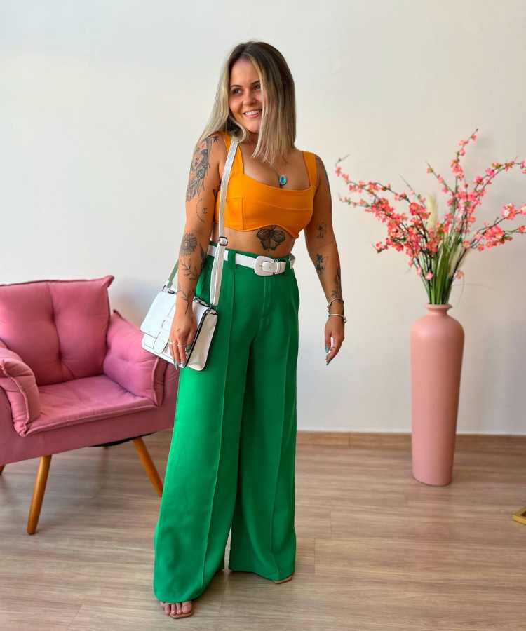 Imagem com fundo de parede, cadeira e vaso de flor. No centro, mulher cropped amarelo, calça verde, cinto branco e bolsa brancausando