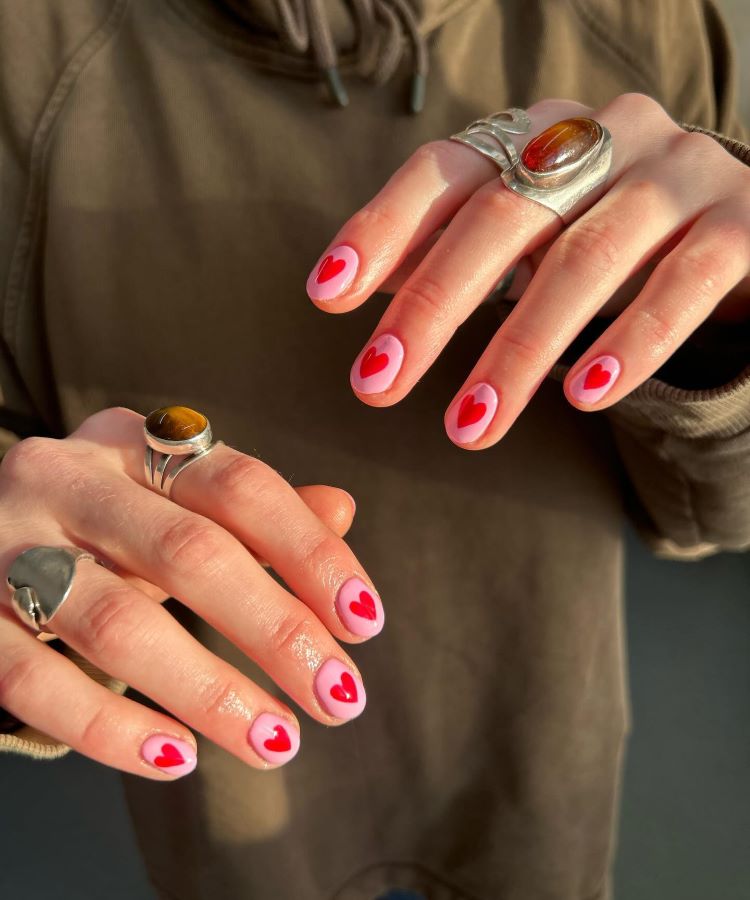 Mãos com unhas pintadas de rosa claro com corações vermelhos no centro. A pessoa usa anéis grandes, incluindo um com pedra tigrada. A luz do sol destaca os detalhes, enquanto o fundo desfocado mostra um moletom marrom.