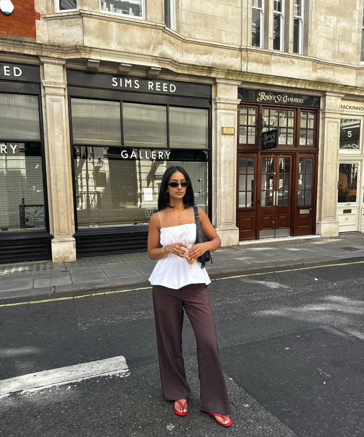 Uma mulher elegante, de óculos de sol, blusa branca com alças finas e calças marrom largas, posa em uma rua. Ela segura uma bolsa preta, um copo com uma bebida e chinelos de dedo vermelhos, em frente a lojas.