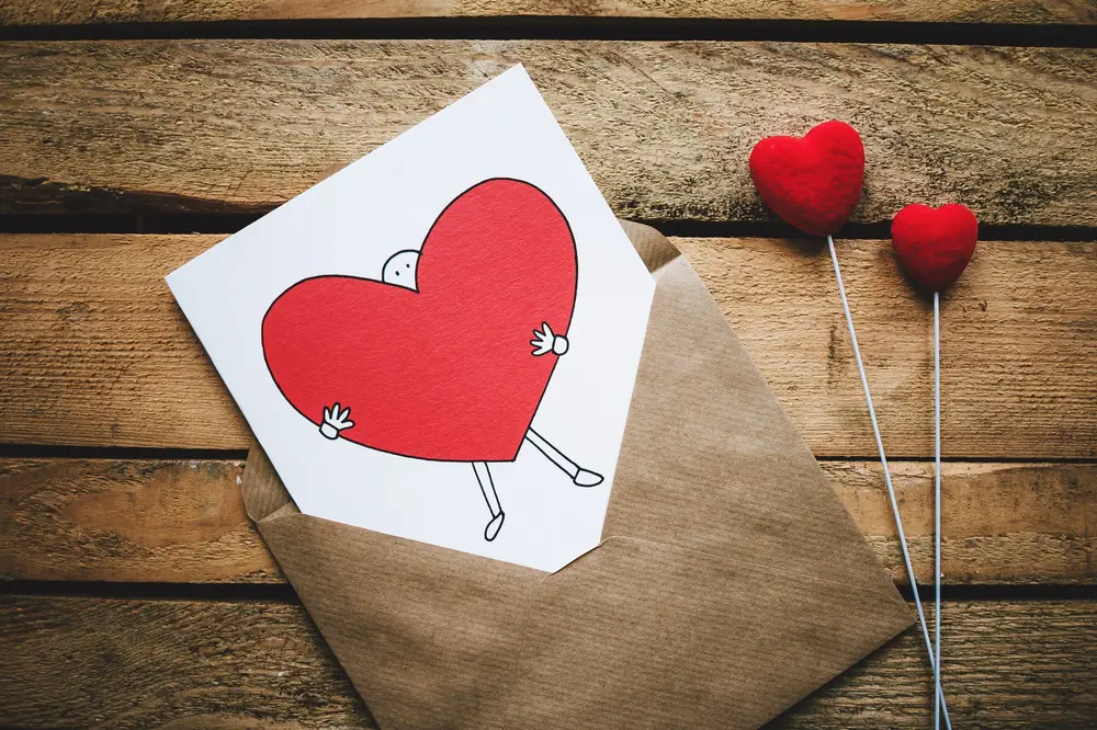 Foto de envelope marrom com cartão branco desenhado com coração e duas hastes com corações vermelhos, tudo sobre superfície de madeira