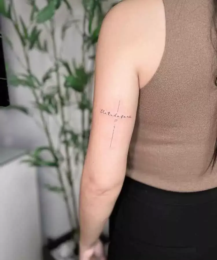 A imagem mostra uma tatuagem delicada na parte posterior do braço de uma mulher. A tatuagem inclui a frase cursiva "ele, do lar" cortada por uma linha vertical, acompanhada dos números "475". A mulher usa uma blusa marrom sem mangas, e o fundo exibe plantas decorativas.