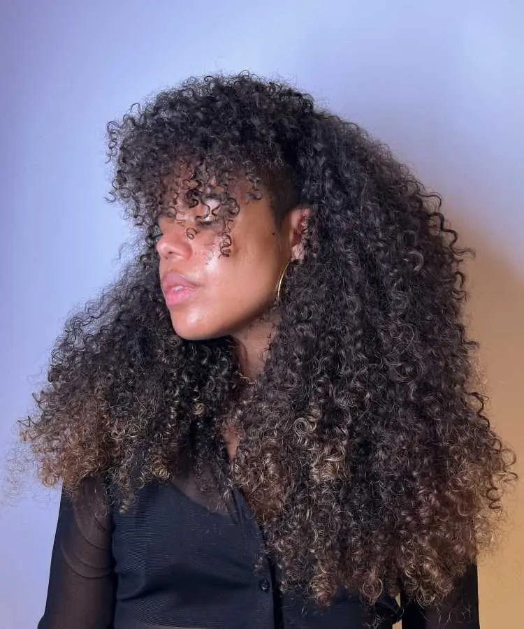 Mulher de perfil com cabelos longos, volumosos e cacheados em tons castanhos, exibindo corte shaggy com franja desfiada. Ela veste blusa preta transparente e brincos dourados, destacando sua expressão confiante contra um fundo neutro com iluminação suave.