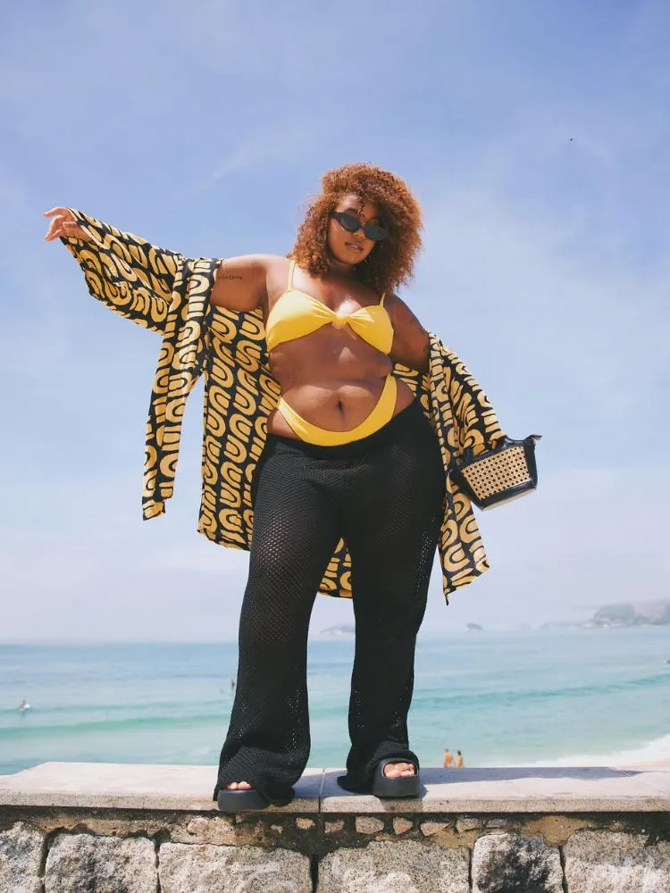 Mari Lobo de biquíni amarelo com camisa amarela e preta, calça preta de crochê, bolsa com detalhe em palha e tamanco preto