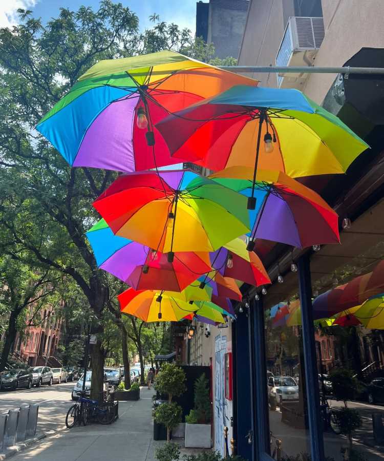 Fileira suspensa de guarda-chuvas coloridos em tons de arco-íris decora a fachada de um prédio. Reflexos nas janelas e luminárias pendentes complementam o cenário. Rua arborizada com bicicleta estacionada e prédios ao fundo cria atmosfera urbana vibrante.
